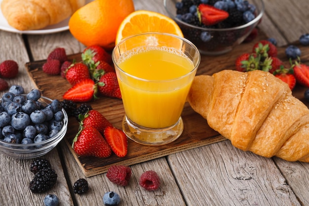 Rijke smakelijke ontbijtachtergrond. Croissants met gouden korst, glas verse jus d'orange en veel zoete biologische bessen op een houten bord over rustieke ondergrond. Heerlijke start van de dag