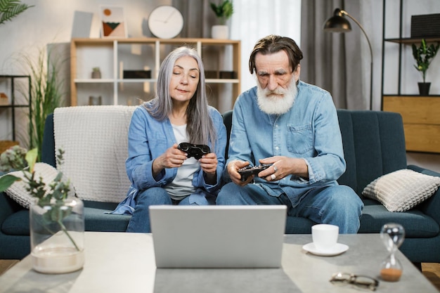 Rijp getrouwd stel met joysticks in handen die videogames spelen terwijl ze op een comfortabele bank zitten