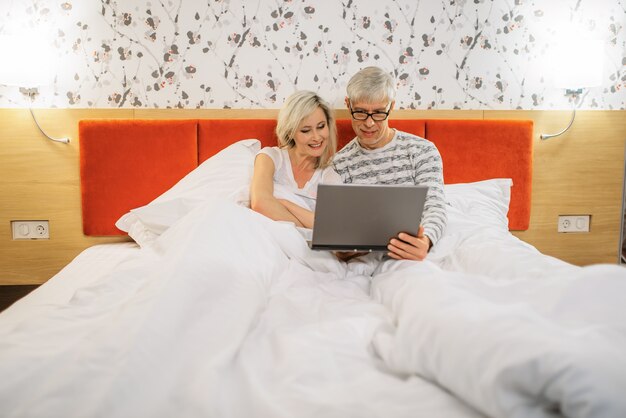 Rijp paar dat op laptop in slaapkamer kijkt alvorens te slapen. man in glazen en vrouw in bed liggen