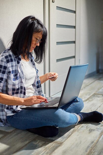 Rijpe aantrekkelijke vrouw met laptopcomputer en creditcard, online winkelen, boekingshotel betalen, online betalen terwijl ze thuis op de vloer zitten.