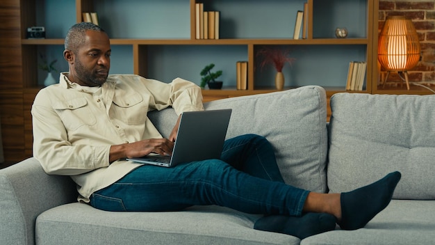 Rijpe Afrikaanse man liggend op de bank ontspannen typen op laptop