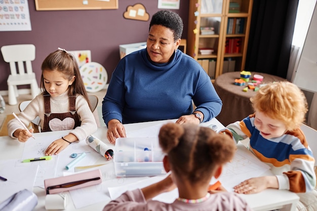 Rijpe Afro-Amerikaanse leraar van de kleuterschool in donkerblauwe pullover die tussen multicultureel zit