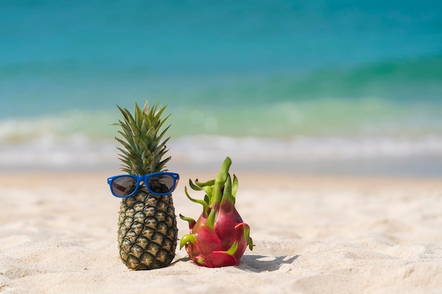 Rijpe ananas in zonnebril en roze drakenfruit op tropisch strand in de buurt van zee