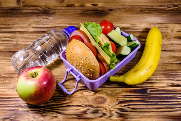 Rijpe appel banaan fles water en lunchbox met hamburger, komkommers en tomaten op houten tafel