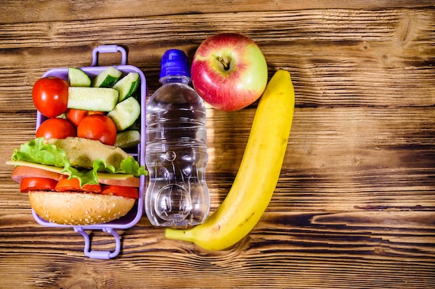 Rijpe appel-banaan fles water en lunchbox met hamburger, komkommers en tomaten op rustieke houten tafel Top view