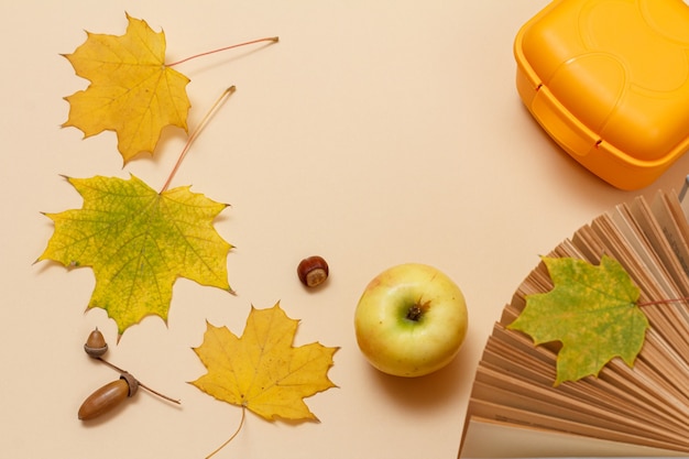 Rijpe appel, een boek, een plastic lunchbox, droge gele esdoornbladeren en eikels op een beige achtergrond. Bovenaanzicht.