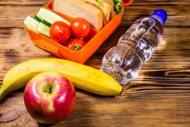 Rijpe appelbanaan fles water en lunchbox met sandwiches, komkommers en tomaten op rustieke houten tafel