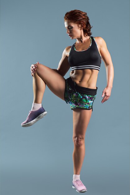 Rijpe atletische roodharige vrouw in sportkleding t-shirt en korte broek fitness oefeningen, grijze studioruimte