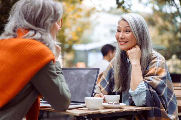 Rijpe aziatische dame en grijsharige vriend met laptop zitten samen op caféterras in de openlucht