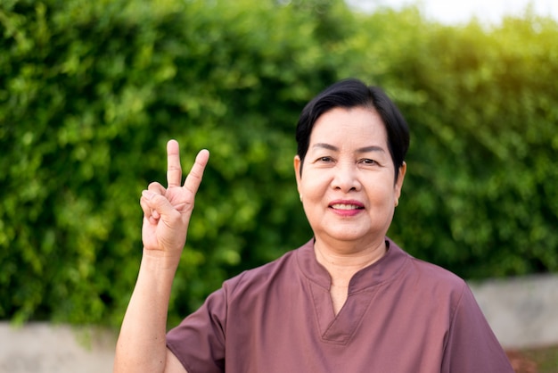 Rijpe Aziatische vrouw die en 2 vingers bevinden zich tonen bij openbaar park, Gelukkig en glimlachend, het Hogere concept van de zorgverzekering