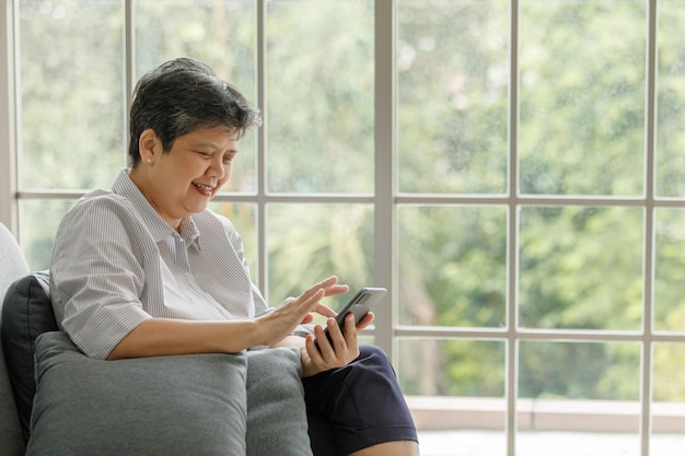 Rijpe Aziatische vrouw lacht en bladert op smartphone terwijl ze in het weekend thuis op de bank bij het raam zit