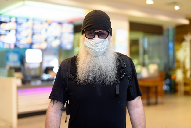Rijpe bebaarde hipster man als backpacker masker en zonnebril dragen in het winkelcentrum