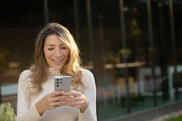 Rijpe blonde vrouw die lacht op haar mobiele telefoonscherm in de stad