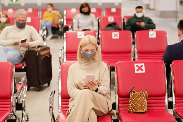 Rijpe blonde vrouwelijke reiziger in vrijetijdskleding en beschermend masker met smartphone tegen andere mensen die wachten op vluchtaankondiging