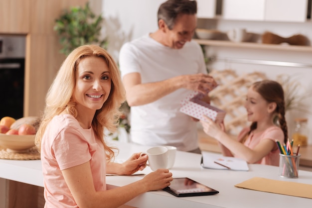 Rijpe charmante positieve moeder genieten van vakantie met het gezin thuis en gadget gebruiken terwijl echtgenoot geschenkdoos geeft aan de dochter