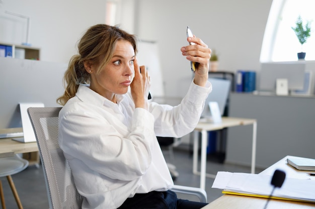 Rijpe elegante vrouw baas met een spiegel corrigeert make-up zittend op de werkplek