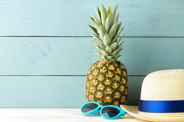 Foto rijpe fruitananas en strandaccessoires, zonnebrillen en hoed op een blauwe houten tafel. zomer.