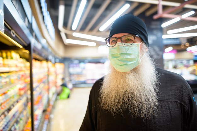 Rijpe gebaarde hipstermens met masker het kopen boodschappen terwijl het handhaven van afstand bij de supermarkt