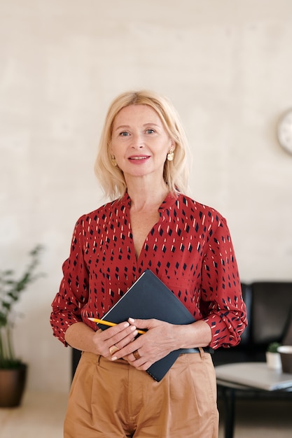 Rijpe gelukkige blonde zakenvrouw in rode blouse en beige broek met notitieboekje en potlood terwijl ze in een kantooromgeving staat