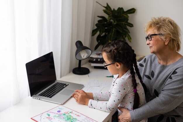 Rijpe grootmoeder helpt kind thuis met huiswerk. Tevreden oude oma helpt haar kleindochter met studeren in de woonkamer. Klein meisje schrijft op notitieboekje met senior leraar naast haar