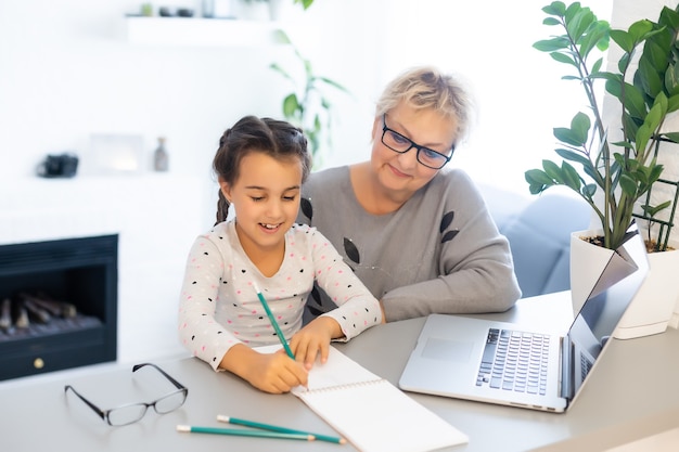 Rijpe grootmoeder helpt kind thuis met huiswerk. Tevreden oude oma helpt haar kleindochter met studeren in de woonkamer. Klein meisje schrijft op notitieboekje met senior leraar naast haar