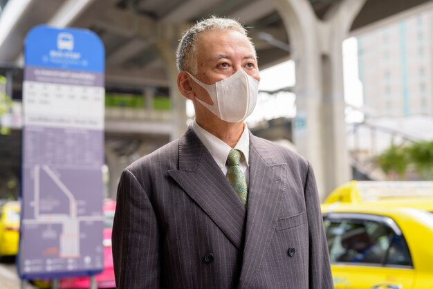 Rijpe Japanse zakenman die met masker bij de bushalte denken