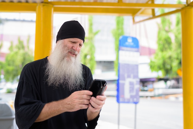 Rijpe knappe bebaarde hipster man met behulp van telefoon zittend bij de bushalte