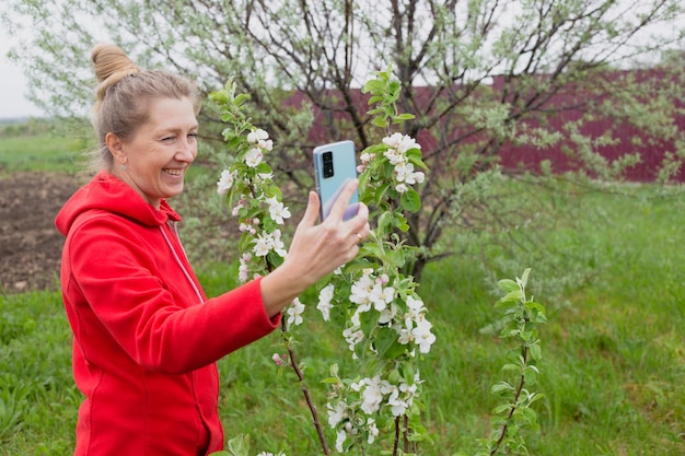 Rijpe lachende vrouw wordt gefotografeerd aan de telefoon met een bloeiende jonge appelboom in haar tuin in het voorjaar