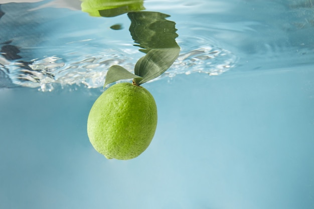 Rijpe limoen met groene bladeren in water geïsoleerd op een blauwe achtergrond, golven op het wateroppervlak. Het concept van een verfrissend zomerdrankje