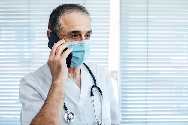 Rijpe mannelijke arts - verpleegster die gezichtsmasker draagt, die op mobiele telefoon naast een het ziekenhuisvenster spreekt. Covid-19 en medicijnconcept