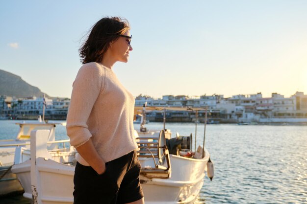 Rijpe mooie gelukkige vrouw in zonnebril wandelen langs de pier, zomer zee vakantie reizen. Zee, afgemeerde boten, bergen, zonsondergang op de achtergrond