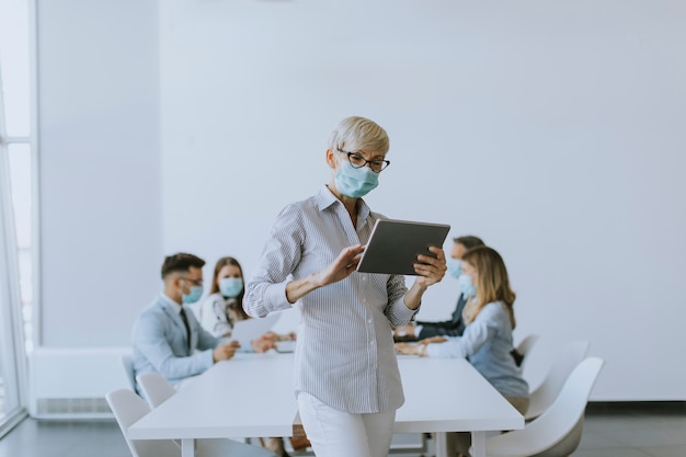 Rijpe onderneemster met beschermend masker die digitale tablet in het bureau gebruiken