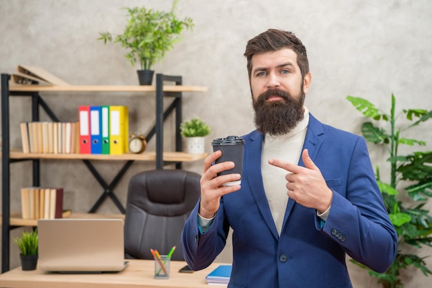 Rijpe ondernemer in jas met koffiepauze puntvinger op kopje tijdens koffiepauze op kantoor