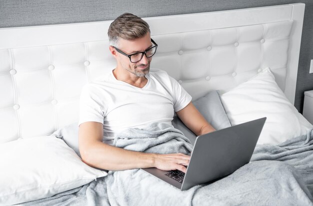 Rijpe online manager in glazen die aan laptop in bed werken