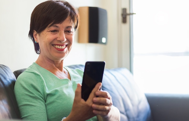 Rijpe oudere vrouw die lacht met behulp van smartphone zittend op de bank thuis