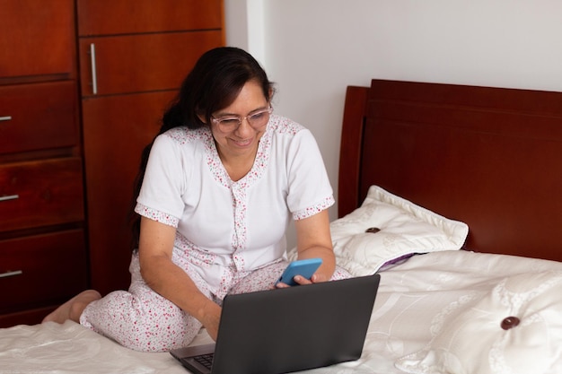 Rijpe, productieve vrouw van 52 jaar die haar laptop en smartphone gebruikt om vanuit bed te werken
