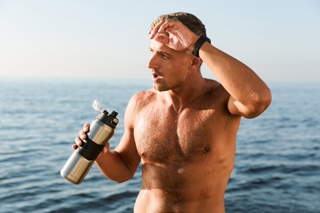 Rijpe sportman die zich met fles water bevindt die op het strand drinkt.