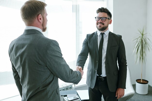 Rijpe succesvolle zakenman in formalwear die zijn zakenpartner tijdens handdruk bekijkt na het onderhandelen en ondertekenen van contract