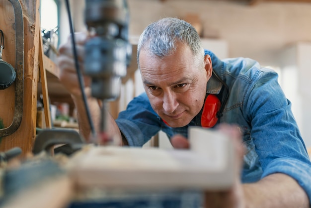 Foto rijpe timmerman die boorpers in workshop met behulp van. hout werken.