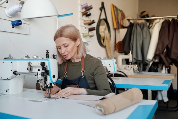 Rijpe vakvrouw in werkkleding die nieuwe leren kleding of andere items naait