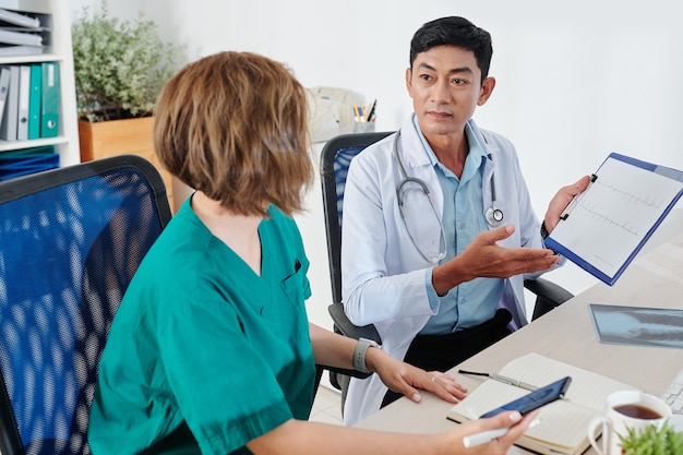 Rijpe Vietnamese cardioloog die het cardiogram van de patiënt laat zien wanneer hij met een collega praat