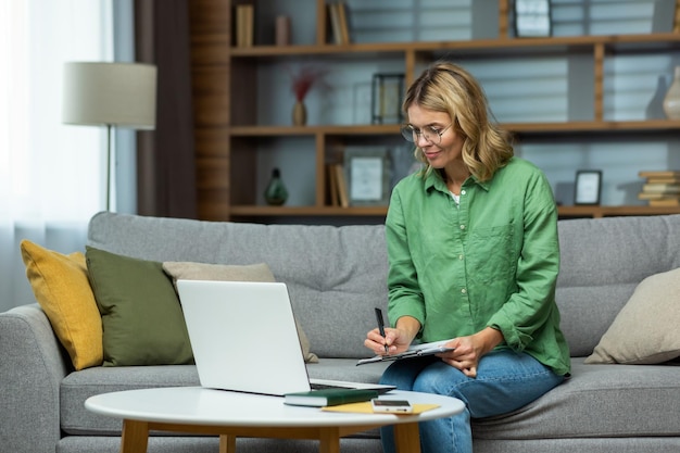 Rijpe vrouw alleen thuis zittend op de bank met laptop rekeningen betalen en werken met documenten