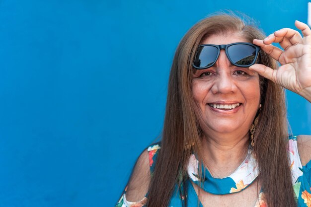 Rijpe vrouw die haar zonnebril steunt. Vrolijke vrouw op blauwe achtergrond.