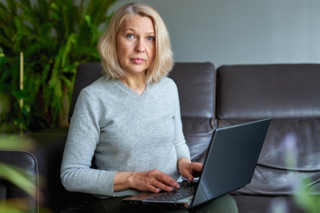 Rijpe vrouw die online zitting aan een bank in een huis werkt