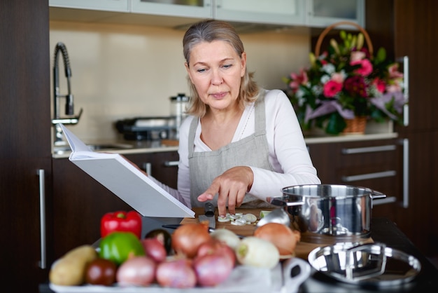 Rijpe vrouw die voedsel in keuken voorbereidt en receptenboek bekijkt