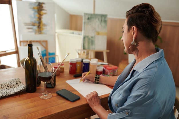 Foto rijpe vrouw die wijn drinkt bij het tekenen van foto's aan een bureau in haar studio