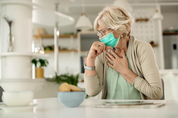 Rijpe vrouw hoest terwijl ze thuis een beschermend gezichtsmasker draagt