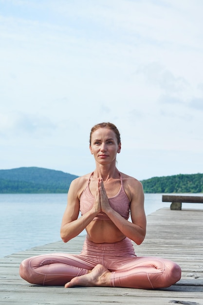 Rijpe vrouw in een roze trainingspak zit in een lotushouding en beoefent yoga in de buurt van de zee Gezond levensstijlconcept