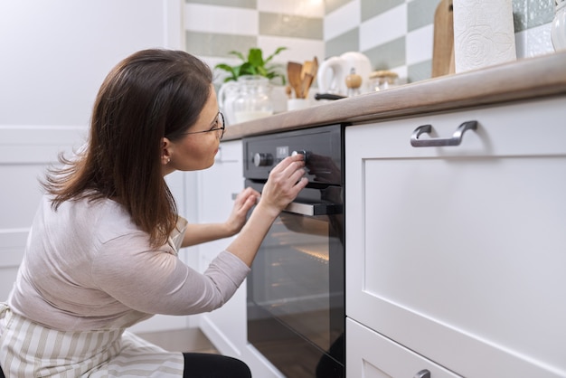 Rijpe vrouw in schort dichtbij de oven in keuken. Huisvrouw zet de instellingen op het fornuis aan, kopieer ruimte