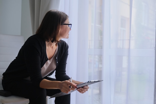 Rijpe vrouw maatschappelijk werker, psycholoog zit bij het raam met klembord.